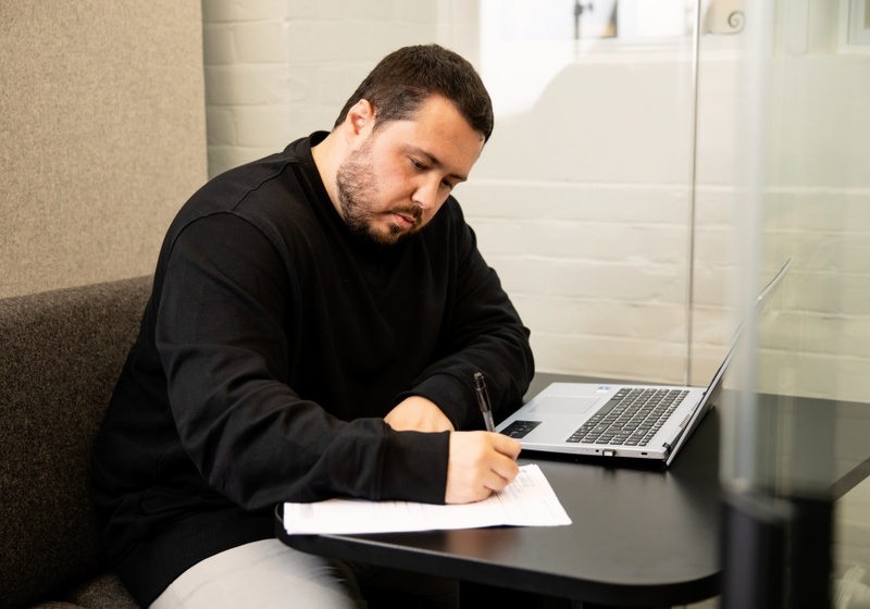 Man in black jumper taking notes at a computer