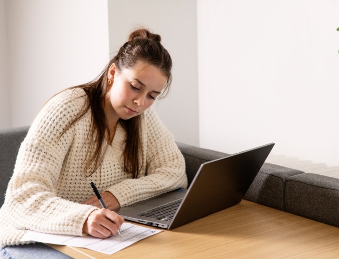 A woman with long hair in a cream jumper. She is taking notes while looking at a laptop.